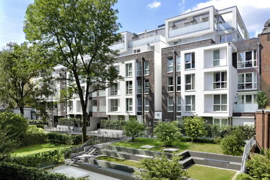 Modern apartment block with large windows and balconies, surrounded by trees and greenery. The landscaped courtyard features tiered grassy areas and hedges, creating a serene and inviting outdoor space under a blue sky.