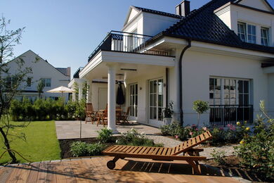Modernes weißes zweistöckiges Haus mit dunklem Dach, großen Fenstern und einem Balkon. Der Garten hinter dem Haus verfügt über eine Terrasse mit Gartenmöbeln, einer Holzliege, grünem Gras und Pflanzen unter einem blauen Himmel.