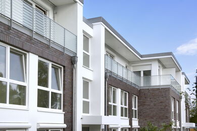 Modern apartment block with large windows, white and dark brick exterior, and balconies with glass balustrades, set against a clear blue sky.