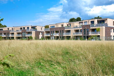Ein modernes, mehrstöckiges Wohnhaus mit Balkonen ist hinter einem Feld mit hohem, gelb-grünem Gras unter einem teilweise bewölkten blauen Himmel zu sehen. Im Hintergrund sind Bäume zu sehen.