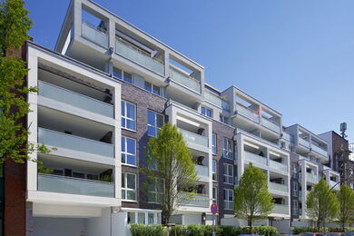 Modern block of flats with multiple storeys, large balconies, and glass balustrades. Trees line the pavement in front of the building under a clear blue sky.
