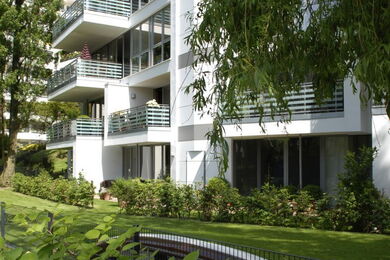 Moderner Wohnblock mit mehreren Balkonen, großen Glasfenstern und weißer Fassade. Das Gebäude ist von grünem Rasen, Sträuchern und Bäumen umgeben, was auf eine ruhige Wohngegend schließen lässt.