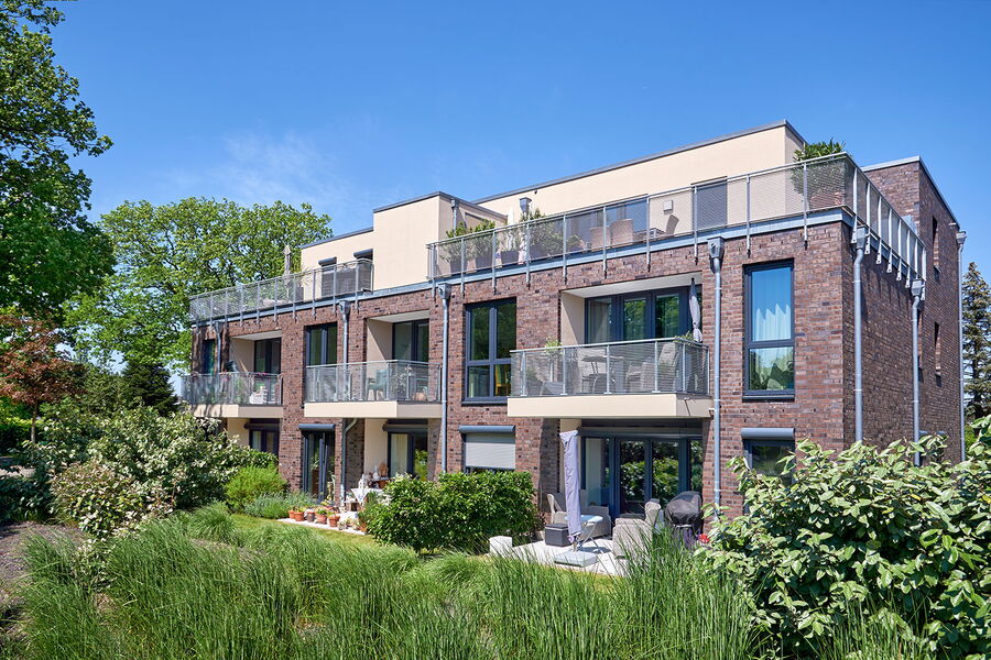 Modernes dreistöckiges Wohnhaus mit großen Balkonen und Backsteinfassade, umgeben von grünen Pflanzen und Bäumen unter einem klaren blauen Himmel. Auf den Balkonen und Terrassen sind Gartenmöbel zu sehen.