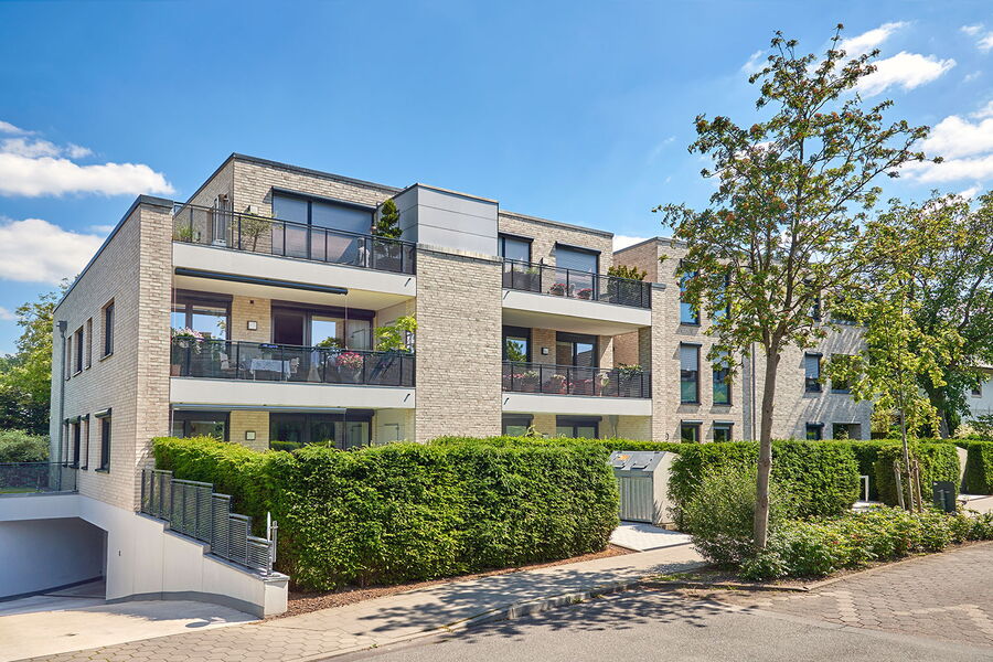 Modernes dreistöckiges Wohnhaus mit Balkonen, großen Fenstern und einer hellen Backsteinfassade, umgeben von grünen Hecken und Bäumen, an einem sonnigen Tag mit blauem Himmel und einigen Wolken.