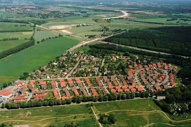 Aerial view of a suburban neighbourhood with red-roofed houses surrounded by green fields, trees, and roads, with a river and distant town visible in the background.