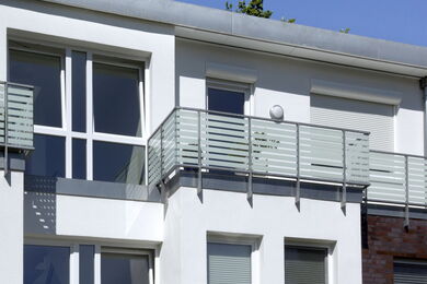 Moderne weiße Gebäudefassade mit großen Fenstern und einem Balkongeländer aus Metall. Einige Fenster haben geschlossene Fensterläden, und der Himmel über dem Gebäude ist klar und blau.