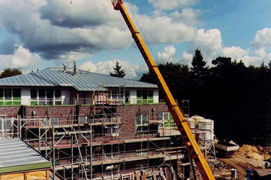 Ein großer gelber Kran hebt Materialien auf einer Baustelle vor einem teilweise errichteten zweistöckigen Gebäude mit Gerüst, umgeben von Bäumen unter einem teilweise bewölkten Himmel.