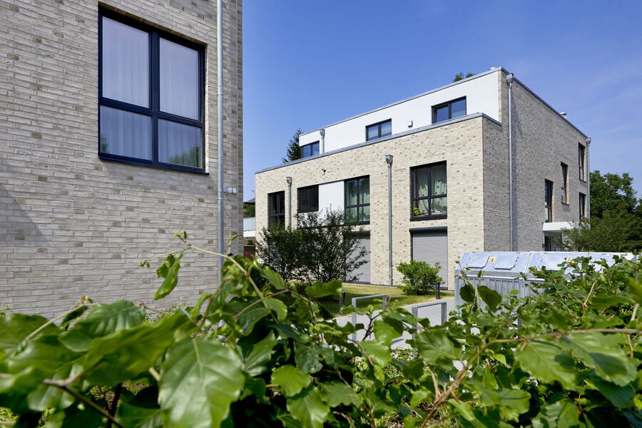 Moderne, helle Backstein-Flachbauten mit großen Fenstern, gesehen hinter grünen Sträuchern und Blättern an einem sonnigen Tag mit strahlend blauem Himmel.