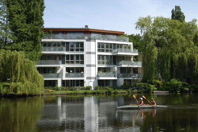 Modern apartment block with large windows overlooks a calm lake, surrounded by greenery and weeping willow trees. Two people in a canoe paddle on the water in front of the block.