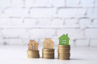Drei kleine hausförmige Ausschnitte sitzen auf Münzstapeln, wobei das Haus ganz rechts grün ist und auf dem höchsten Stapel liegt. Der Hintergrund ist eine weiße Backsteinmauer, die Immobilieninvestitionen oder Ersparnisse symbolisiert.